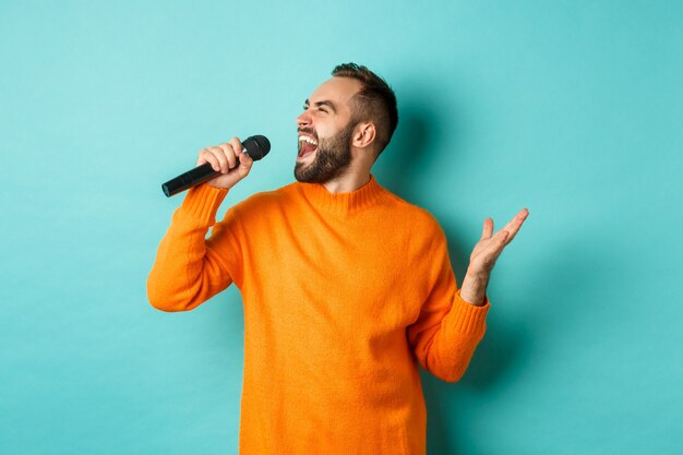 Apuesto hombre adulto realiza una canción, cantando en el micrófono, de pie contra la pared turquesa