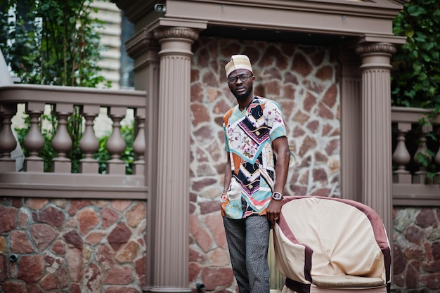 Apuesto hombre afroamericano con gorra de ropa tradicional y anteojos en la ciudad moderna de pie cerca de la silla