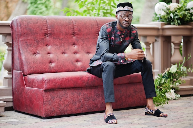 Foto gratuita apuesto hombre afroamericano con gorra de ropa tradicional y anteojos en la ciudad moderna sentado en el sofá