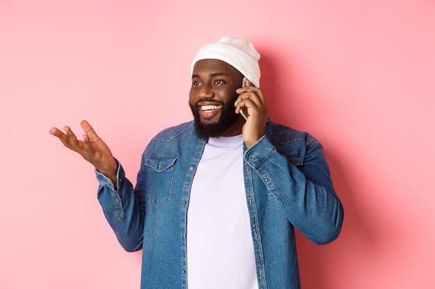 Apuesto hombre afroamericano moderno hablando por teléfono móvil, sonriendo y discutiendo algo, de pie sobre fondo rosa