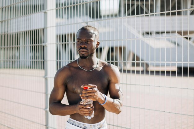 Apuesto hombre afroamericano con pecho desnudo posa con una botella de agua