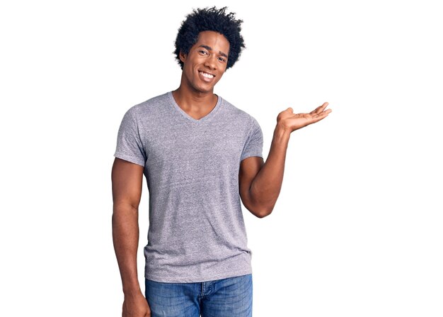 Un apuesto hombre afroamericano con el pelo afro vestido con ropa informal sonriendo alegremente presentando y señalando con la palma de la mano mirando la cámara.