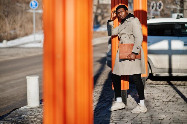 Apuesto hombre afroamericano posando afuera con sombrero negro y abrigo beige con carpeta en la mano