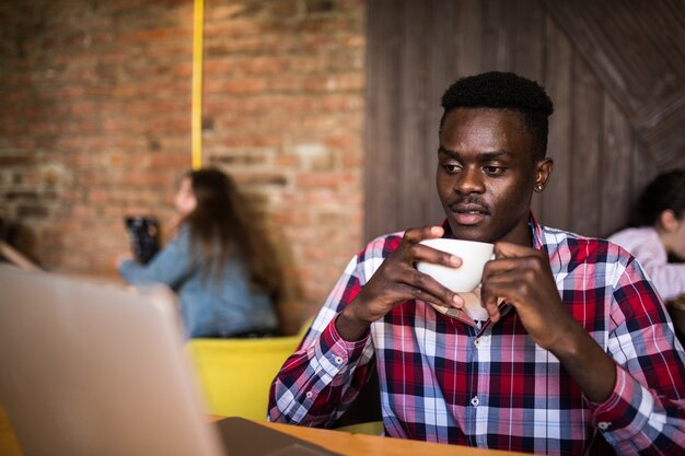 Apuesto hombre afroamericano en ropa casual sosteniendo una taza de café y usando laptop.