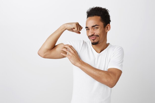 Apuesto hombre afroamericano seguro y fuerte flexionando bíceps, entrenamiento en el gimnasio, mirando descarado
