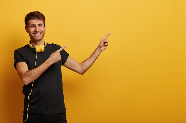 Apuesto hombre alegre apunta al espacio de la copia, vestido con ropa negra, usa auriculares, sonríe con dientes, demuestra publicidad, aislado sobre fondo amarillo