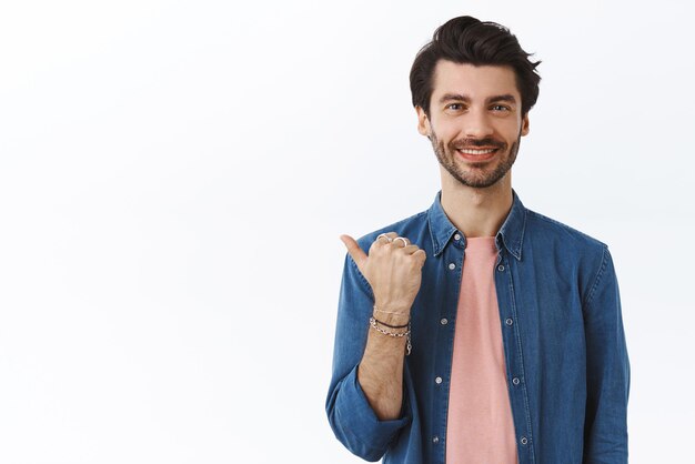 Un apuesto hombre alegre que le muestra el lugar donde puede encontrar las mejores opciones para los regalos de Navidad que señala con el pulgar a la izquierda sonriendo alegremente da consejos o sugerencias para promover el fondo blanco del producto