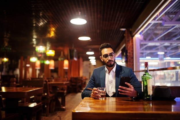 Foto gratuita un apuesto hombre árabe bien vestido con un vaso de whisky y un cigarro sostiene un teléfono móvil en el pub