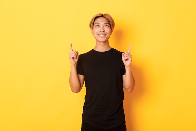 Apuesto hombre asiático sonriente en camiseta negra apuntando con el dedo hacia arriba, pared amarilla.