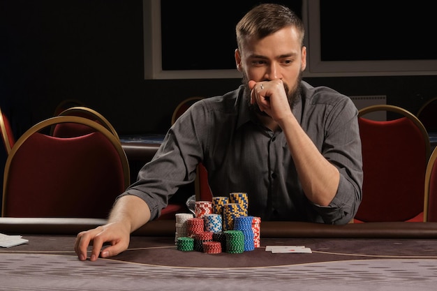 Foto gratuita un apuesto hombre barbudo con una camisa gris está jugando al póquer sentado en la mesa del casino. está haciendo apuestas esperando una gran victoria. juegos de azar por dinero. juegos de fortuna.