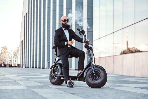 Un apuesto hombre barbudo está haciendo un breve descanso mientras fuma un vaporizador. El hombre está sentado en un scooter eléctrico.