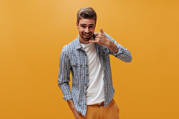 Apuesto hombre barbudo en pantalones brillantes camiseta blanca y camisa a cuadros sonríe mira a la cámara y muestra señal de llamada sobre fondo naranja