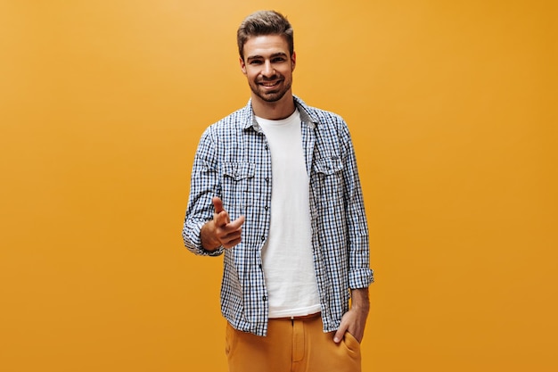 Foto gratuita un apuesto hombre barbudo con pantalones naranja, camiseta blanca y camisa a cuadros sonríe sinceramente y señala a la cámara con fondo naranja