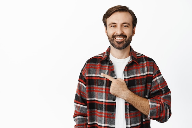 Un apuesto hombre barbudo sonriente de 30 años señalando con el dedo a la izquierda mostrando un anuncio de pie sobre fondo blanco