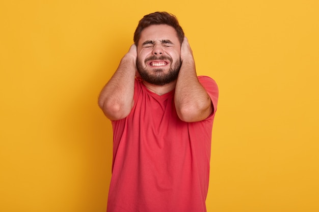 apuesto hombre barbudo vistiendo camiseta redcasual, hombre atractivo posando con los ojos y oídos cerrados, escuchar ruidos fuertes, chico de pie aislado en amarillo. Concepto de personas