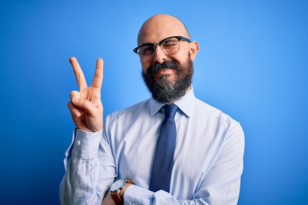 Apuesto hombre calvo de negocios con barba con elegante corbata y anteojos sobre fondo azul sonriendo con cara feliz guiñando un ojo a la cámara haciendo el signo de la victoria Número dos