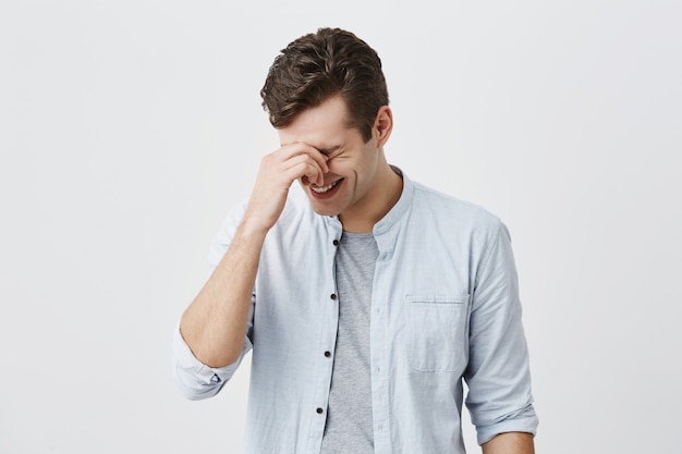 Apuesto hombre caucásico guapo con corte de pelo moderno vestido con camisa azul, sonriendo, riéndose de la broma de un amigo o historia divertida, tocando el puente de la nariz. Emociones positivas y concepto de reacción.