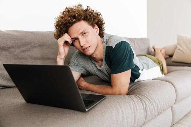 Apuesto hombre concentrado se encuentra en el sofá usando laptop