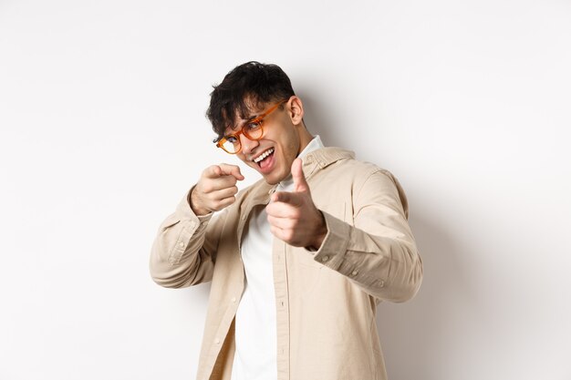 Apuesto hombre divertido con gafas sonriendo, señalando con el dedo a la cámara, eligiendo o invitándote, de pie sobre fondo blanco.