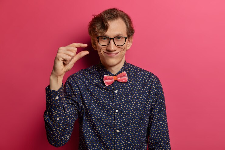 Foto gratuito apuesto hombre empollón positivo hace un signo de mano de tamaño pequeño con los dedos, mide algo pequeño, explica cuánto necesita algo, vestido formalmente, aislado en una pared rosa. muy poco