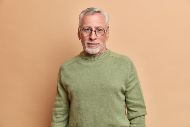 Apuesto hombre europeo barbudo con mirada curiosa usa anteojos y el jersey básico mira directamente al frente posa contra la pared beige