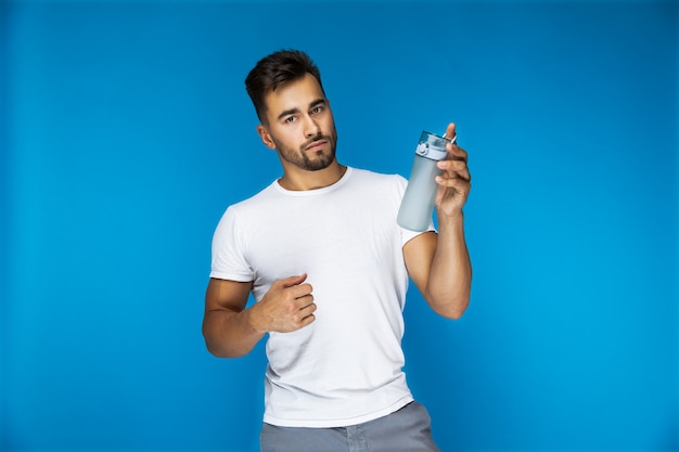 Apuesto hombre europeo en camiseta blanca sobre fondo azul está sosteniendo una botella deportiva en una mano