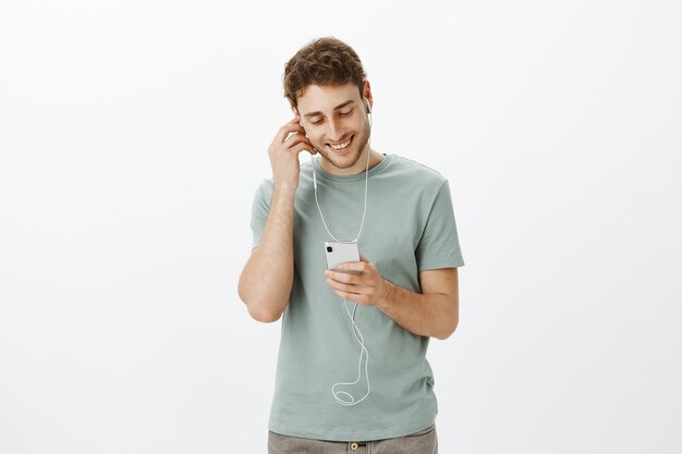 Apuesto hombre europeo despreocupado con cabello rubio y cerdas, sosteniendo la mano en el auricular y sonriendo ampliamente en la pantalla del teléfono inteligente, disfrutando de la nueva canción del cantante favorito