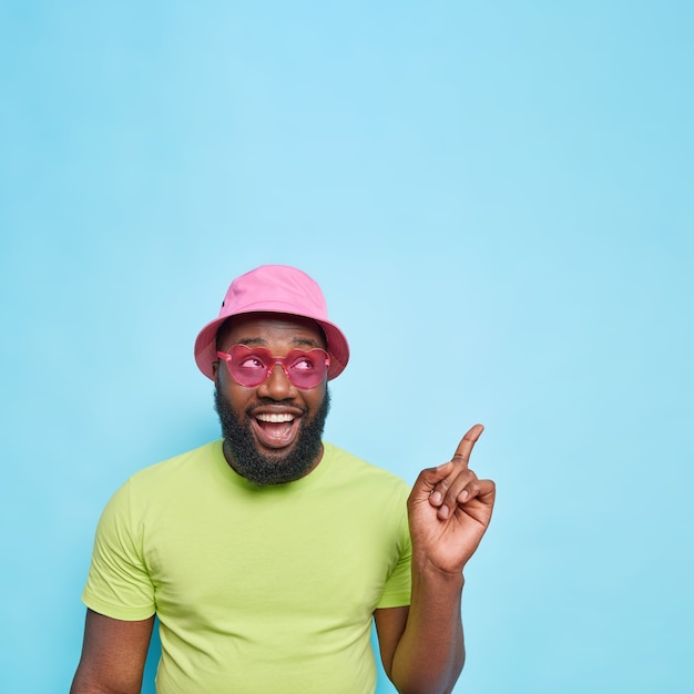 Apuesto hombre feliz con barba gruesa en la esquina superior derecha vestido con ropa de verano, gafas de sol rosa de moda, muestra espacio de copia para su anuncio aislado en la pared azul