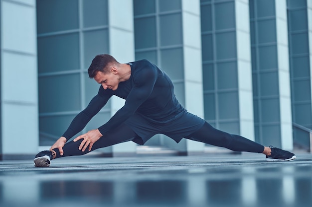 Un apuesto hombre de fitness con ropa deportiva, haciendo estiramientos mientras se prepara para un ejercicio serio en la ciudad moderna contra un rascacielos. Concepto de estilo de vida saludable.