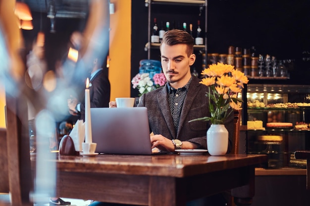 Un apuesto hombre independiente con elegante barba y cabello vestido con un traje negro que trabaja en una laptop mientras está sentado en un café.