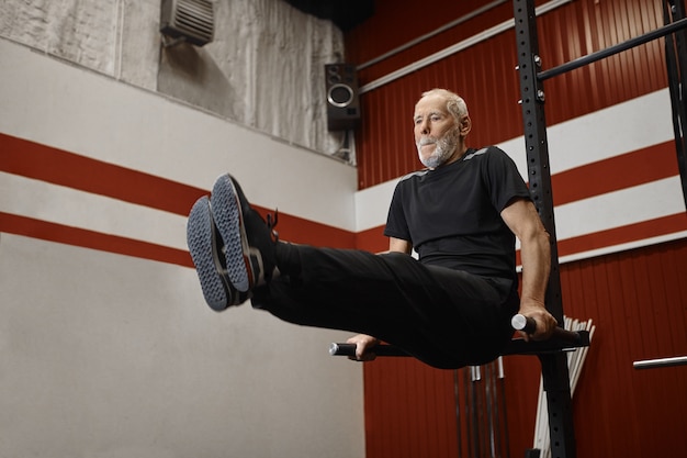 Apuesto hombre jubilado de usnhaven en ropa deportiva elegante haciendo entrenamiento de crossfit en el gimnasio, levantando las piernas mientras hace ejercicio en la barra de tracción, fortaleciendo los abdominales. concepto de fitness, deportes y jubilación.