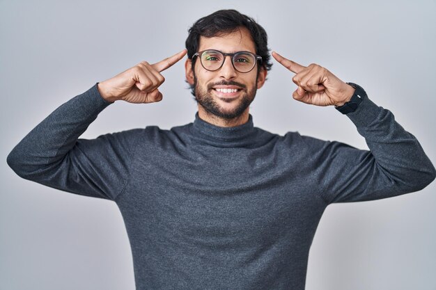 Apuesto hombre latino de pie sobre un fondo aislado sonriendo apuntando a la cabeza con el dedo de ambas manos, gran idea o pensamiento, buena memoria