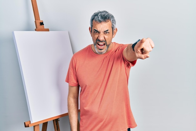 Foto gratuita apuesto hombre de mediana edad con cabello gris de pie junto al caballete del pintor señalando disgustado y frustrado a la cámara, enojado y furioso contigo