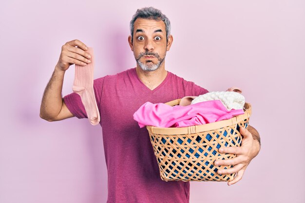 Apuesto hombre de mediana edad con cabello gris sosteniendo una cesta de ropa sucia y calcetín sucio hinchando las mejillas con cara graciosa boca inflada con aire atrapando aire