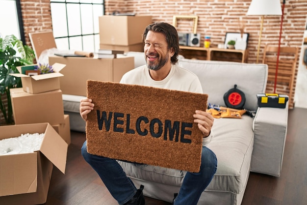 Foto gratuita apuesto hombre de mediana edad con felpudo de bienvenida en el nuevo hogar guiñando un ojo mirando a la cámara con expresión sexy cara alegre y feliz