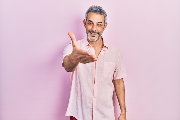 Un apuesto hombre de mediana edad con el pelo gris y un pantalones casual sonriendo amablemente ofreciendo un apretón de manos como saludo y bienvenida. Negocio exitoso.