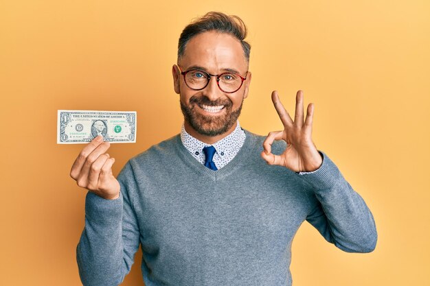 Un apuesto hombre de mediana edad sosteniendo un billete de 1 dólar haciendo un buen signo con los dedos sonriendo amigablemente haciendo un gesto excelente