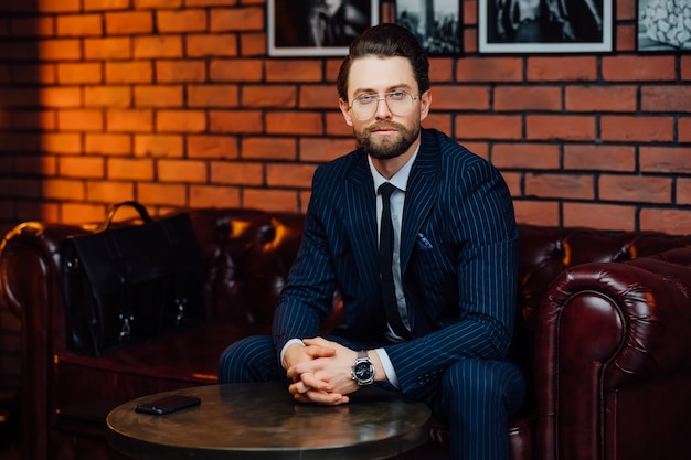 Apuesto hombre de negocios con anteojos de moda y elegante traje sentado en el sofá en el estudio moderno.