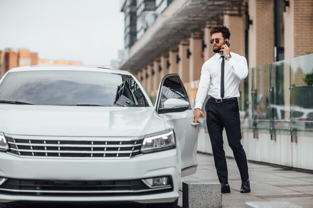 Apuesto hombre de negocios barbudo con camisa blanca, hablando por teléfono y entrando en su automóvil mientras está parado al aire libre en las calles de la ciudad cerca del moderno centro de oficinas