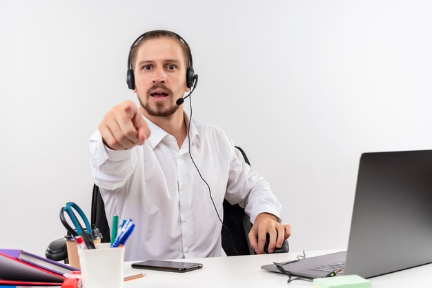 Apuesto hombre de negocios en camisa blanca y auriculares con un micrófono apuntando con el dedo a la cámara mirando confundido sentado en la mesa en offise sobre fondo blanco.