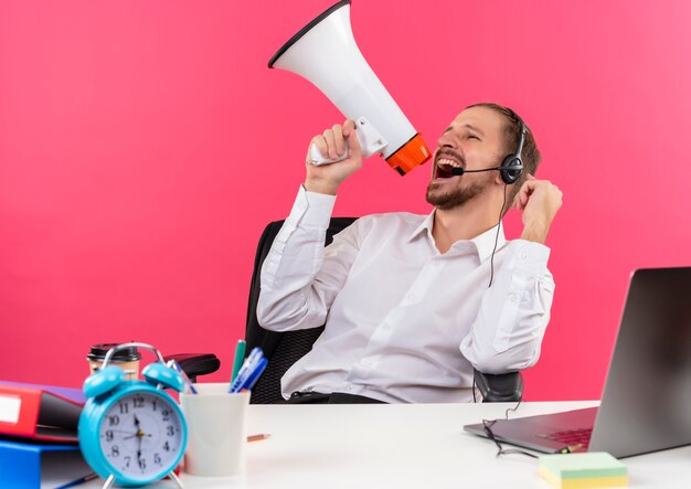 Apuesto hombre de negocios en camisa blanca y auriculares con un micrófono gritando al megáfono apretando el puño loco feliz sentado en la mesa en offise sobre fondo rosa