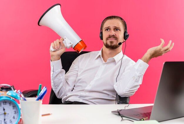 Apuesto hombre de negocios en camisa blanca y auriculares con un micrófono con megáfono mirando confundido sentado en la mesa en offise sobre fondo rosa