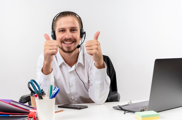 Apuesto hombre de negocios con camisa blanca y auriculares con un micrófono mirando a la cámara sonriendo mostrando los pulgares para arriba sentado en la mesa en offise sobre fondo blanco.