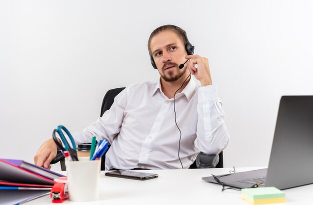 Apuesto hombre de negocios en camisa blanca y auriculares con un micrófono mirando a un lado con cara seria sentado en la mesa en offise sobre fondo blanco.