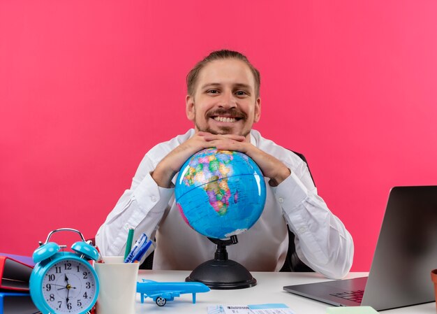 Apuesto hombre de negocios en camisa blanca con globo mirando a la cámara con una sonrisa en la cara sentado en la mesa en offise sobre fondo rosa