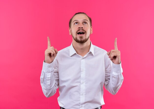 Apuesto hombre de negocios con camisa blanca mirando hacia arriba apuntando con el dedo índice hacia arriba feliz y positivo de pie sobre fondo rosa