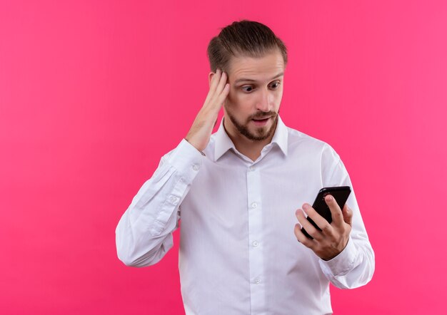 Apuesto hombre de negocios con camisa blanca mirando la pantalla de su teléfono inteligente sorprendido y asombrado de pie sobre fondo rosa