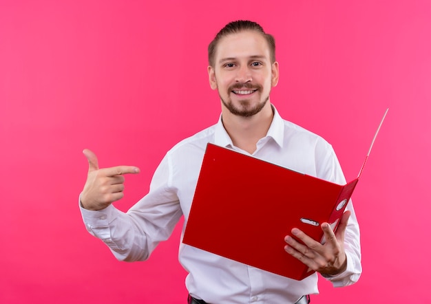 Apuesto hombre de negocios en camisa blanca sosteniendo la carpeta abierta mirando a la cámara apuntando con el dedo sonriendo de pie sobre fondo rosa