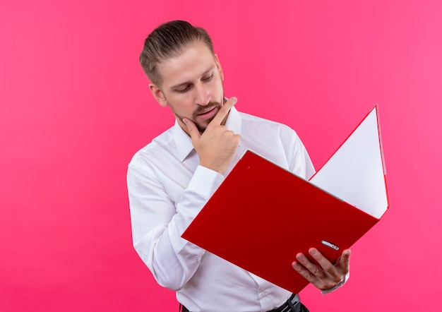 Apuesto hombre de negocios en camisa blanca sosteniendo la carpeta mirándola con cara seria de pie sobre fondo rosa