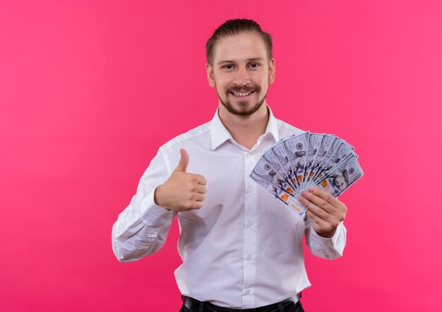 Apuesto hombre de negocios en camisa blanca sosteniendo dinero en efectivo mirando a la cámara sonriendo alegremente mostrando los pulgares para arriba sobre fondo rosa